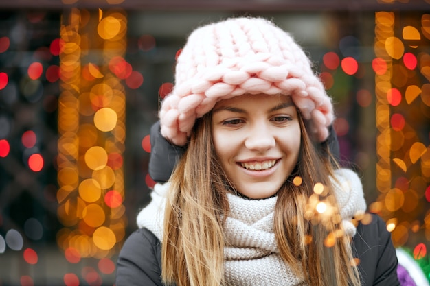 Attraente ragazza bionda sorridente che indossa abiti invernali che celebrano il Natale alle ghirlande sfondo sfocato. Spazio per il testo