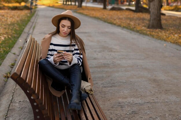 Attraente ragazza bruna allegra in trench con caffè per andare felicemente utilizzando il cellulare all'aperto