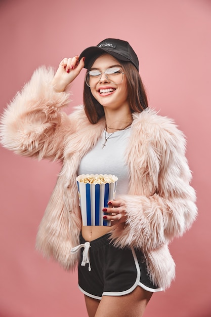 Attraente ragazza bruna in finta pelliccia rosa con popcorn in mano indossando pantaloncini neri top bianco ...