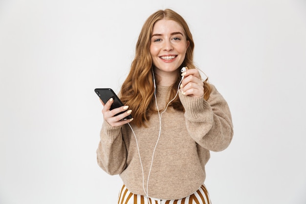 Attraente ragazza che indossa un maglione in piedi isolato su un muro bianco, ascoltando musica con gli auricolari, tenendo il telefono cellulare
