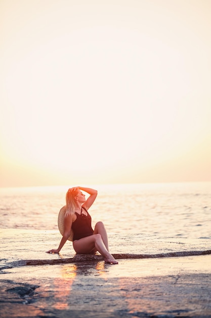 Attraente ragazza con i capelli lunghi pone sulla spiaggia Indossa un costume da bagno nero Luce dorata del tramonto