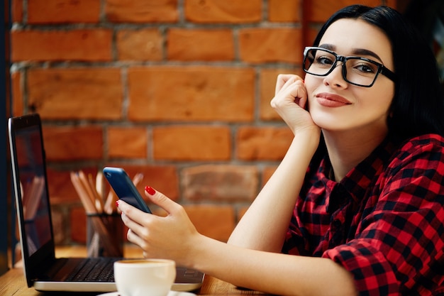 Attraente ragazza con i capelli neri che indossa occhiali da vista seduto in caffè con laptop, telefono cellulare e tazza di caffè, concetto di freelance, ritratto, copia spazio, indossa la camicia rossa.