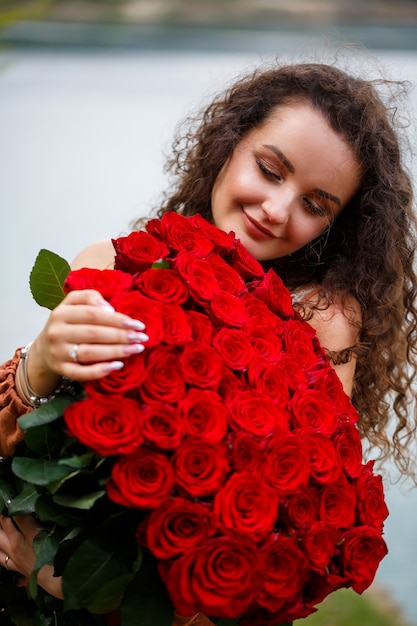 Attraente ragazza con i capelli ricci e un sorriso sul viso con un enorme mazzo di rose rosse su uno sfondo di lago blu. Calda giornata estiva, giovane donna felice, emozioni di gioia