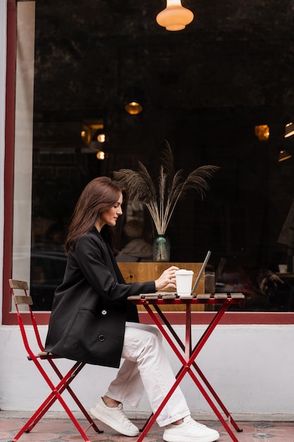 Attraente ragazza con laptop e tazza di caffè è seduta fuori dal bar Giovane donna d'affari con laptop lavora da remoto all'aperto e beve caffè