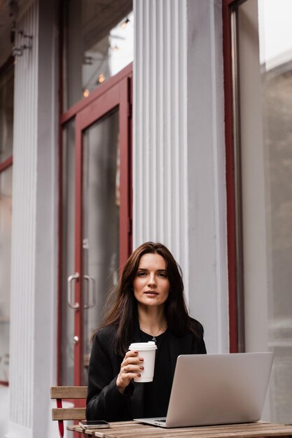 Attraente ragazza con laptop e tazza di caffè è seduta fuori dal caffè Giovane donna d'affari con laptop lavora da remoto all'aperto e beve caffè