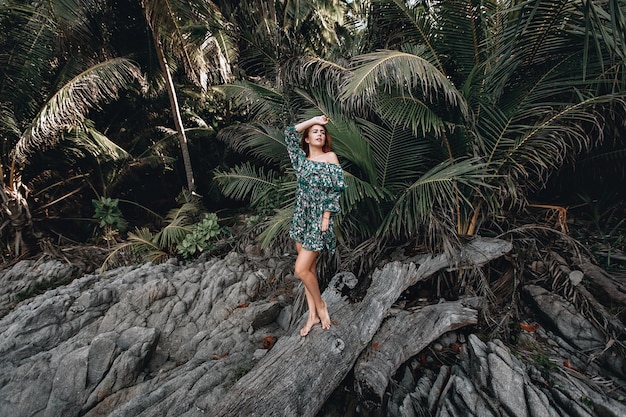 Attraente ragazza dai capelli rossi in un vestito con stampa floreale in posa vicino a grandi palme e in piedi su un vecchio albero. Phuket. Tailandia