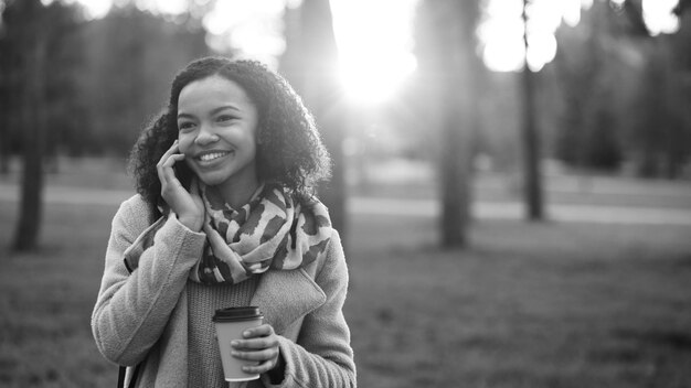 Attraente ragazza di razza mista che parla smartphone e beve caffè nel parco cittadino con borse