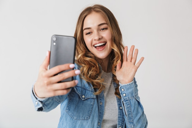 Attraente ragazza eccitata in piedi isolata su bianco, facendo un selfie, salutando