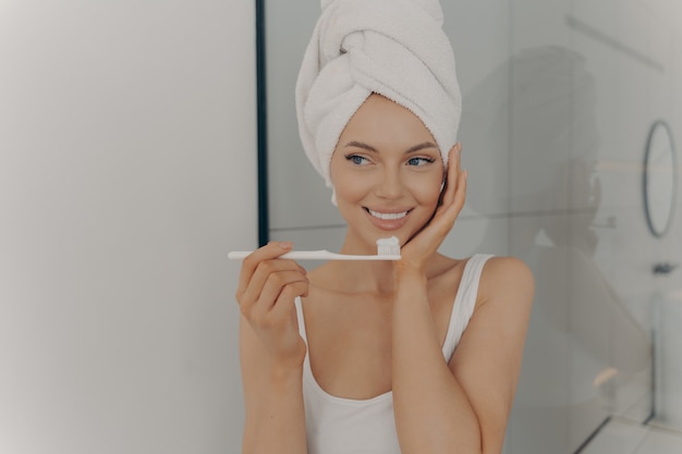 Attraente ragazza felice con un sorriso ampio bianco perfetto che si lava i denti in bagno davanti alla telecamera dopo aver fatto la doccia mattutina. Concetto di salute e igiene dentale