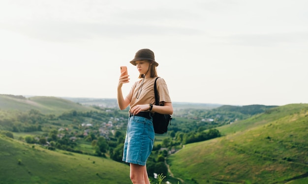 Attraente ragazza hipster in abiti eleganti si trova in cima a una montagna a Tovtry e utilizza uno smartphone Ragazza turista si trova su una roccia nel bellissimo paesaggio ucraino e scatta foto su uno smartphone