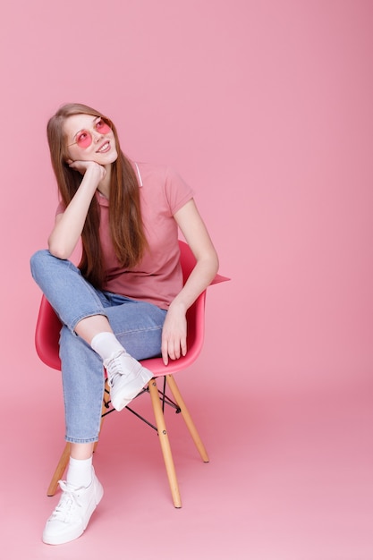 Attraente ragazza in occhiali rosa si siede e sorride su sfondo rosa