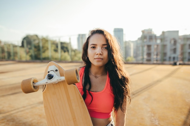 attraente ragazza ispanica in abiti casual in piedi per strada con longboard in mano