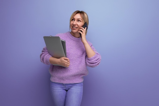 attraente, sicura, intelligente, bionda, donna millenaria che tiene in mano un portatile per il lavoro e parla al telefono sullo sfondo blu