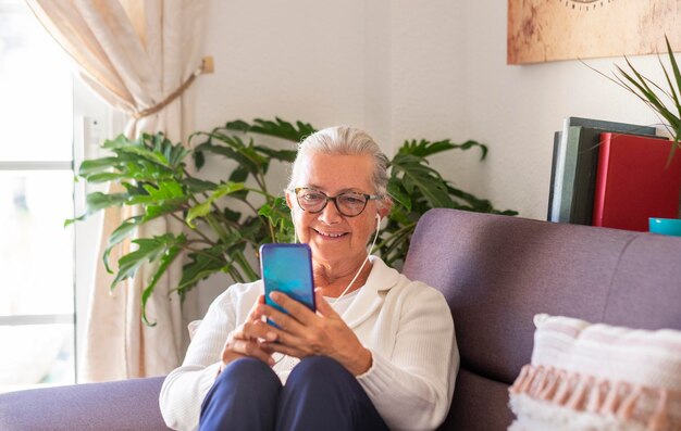 Attraente signora anziana seduta sul divano a casa usando il suo smartphone per chattare con gli amici o la famiglia donna capelli bianchi occhiali che indossano auricolari persone sociali e tecnologiche attive