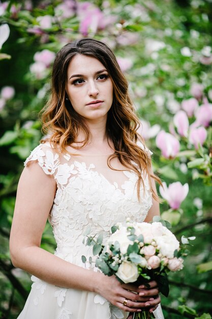 Attraente sposa con un bouquet da sposa in piedi su sfondo di fiori viola di magnolia e verdi. natura. all'aperto. guardando lateralmente. mezza lunghezza superiore.