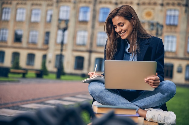 Attraente studentessa bruna seduta con il suo laptop sulle ginocchia mentre studia sulla panchina fuori