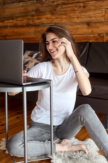 Attraente studentessa sorridente felice con chat video seduto sul pavimento, utilizzando il computer portatile. La donna graziosa gode di guardare il webinar educativo sul laptop.