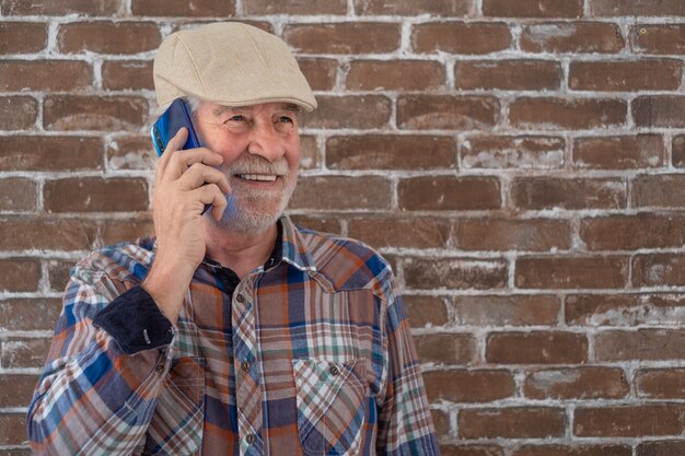 Attraente uomo anziano abbigliamento casual in piedi contro un muro di mattoni utilizzando il telefono e sorridente.