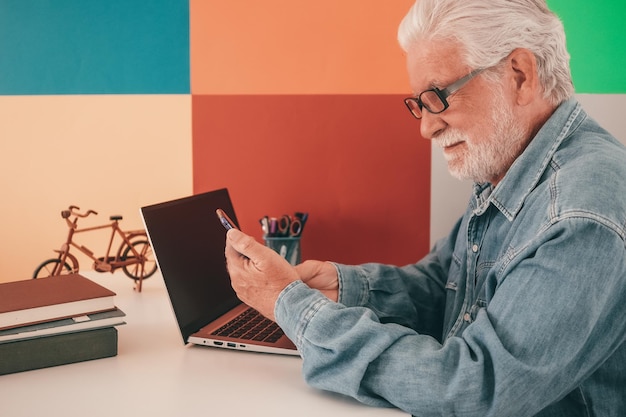 Attraente uomo anziano in camicia di jeans e occhiali seduto alla scrivania dell'ufficio utilizzando lo smartphone