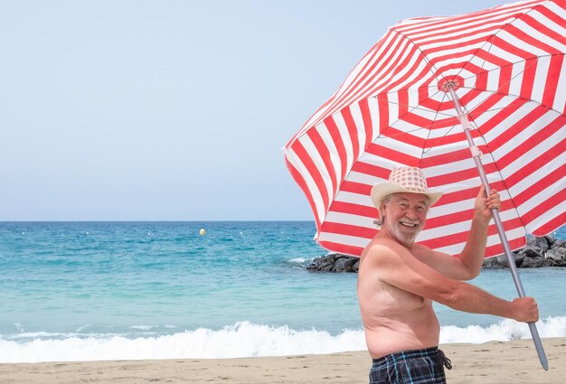 Attraente uomo anziano sorridente con barba e cappello in piedi sulla spiaggia con un ombrello bianco e rosso in mano godendosi l'orizzonte delle vacanze e della libertà sull'acqua