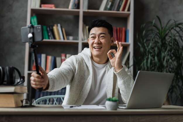 Attraente uomo asiatico in abiti casual prendendo selfie sul suo smartphone utilizzando il bastone che mostra il pollice in su