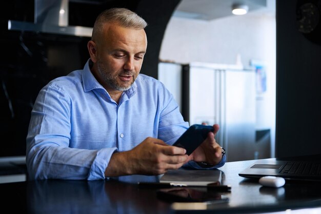 Attraente uomo d'affari di mezza età fiducioso, imprenditore di successo in camicia casual blu sorride usando il telefono cellulare seduto al tavolo in un elegante interno di casa