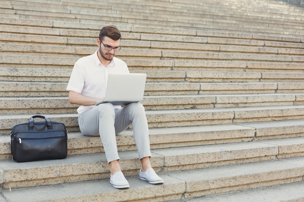 Attraente uomo d'affari premuroso con gli occhiali sta lavorando sul suo computer portatile mentre è seduto su scale di pietra accanto alla sua valigetta, copia spazio. Tecnologia e concetto di business