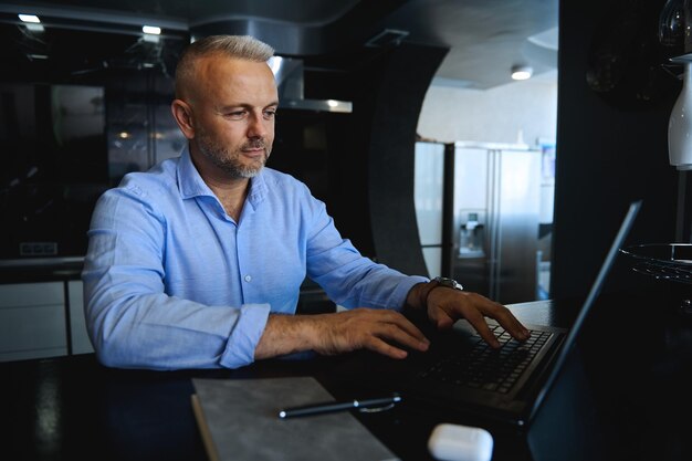 Attraente uomo europeo di mezza età sicuro di sé in camicia casual digitando il testo sulla tastiera del laptop, pianificando progetti, navigando su siti Web mentre si lavora online a casa con interni eleganti