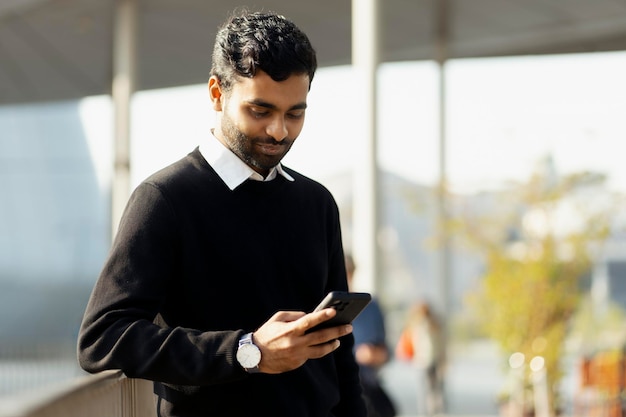 Attraente uomo indiano che utilizza il telefono cellulare leggendo messaggi di testo, comunicazione online per strada