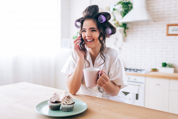 Attrarre la bella casalinga femminile che parla sul telefono. Tenendo la tazza in mano. Pancakes su piastra. Pettegolezzo. Bruna con bigodini nei capelli. Vita spensierata senza lavoro. A casa da solo.