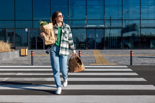 Attraversamento pedonale della donna la strada da un centro commerciale che tiene i sacchetti di carta per lo shopping con generi alimentari