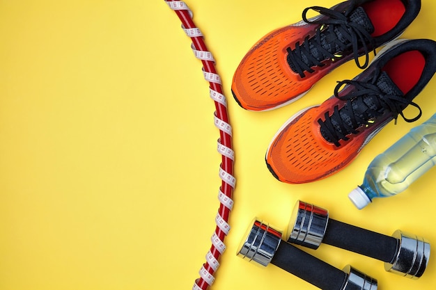 Attrezzatura per esercizi. Sneakers sportive arancioni, cerchio da ginnastica avvolto in un nastro di misurazione con un metro, manubri e una bottiglia d'acqua su sfondo giallo. Esercizi dimagranti e sani per il corpo