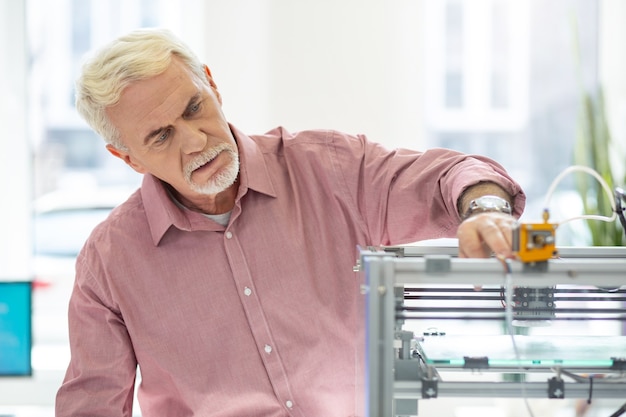 Attrezzature all'avanguardia. Affascinante uomo anziano che utilizza una stampante 3D in ufficio e controlla il suo lavoro