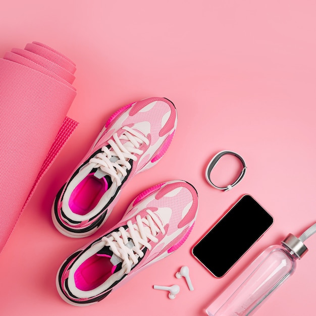 Attrezzature sportive e scarpe da ginnastica su sfondo rosa. Smartphone, braccialetto fitness, tappetino yoga e borraccia. App per l'allenamento indoor. Programma di fitness online. Allenamento online a casa.