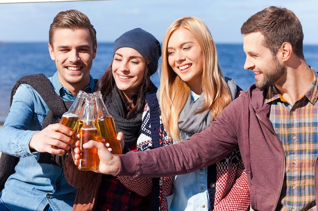 Auguri all'amicizia! Quattro giovani felici che esultano con la birra e sorridono mentre si legano l'un l'altro