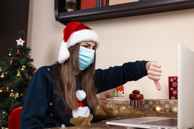 Auguri di Natale in linea. ragazza in cappello di Babbo Natale in colloqui maschera medica Dà un regalo utilizzando laptop per videochiamata amici e genitori. La stanza è addobbata a festa. Natale durante il coronavirus.