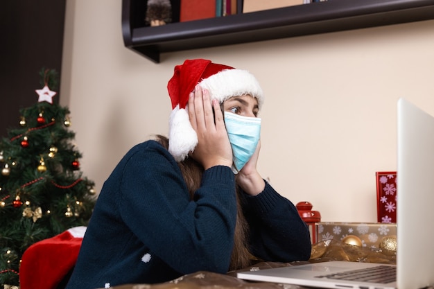 Auguri di Natale in linea. ragazza in cappello di Babbo Natale in colloqui maschera medica Dà un regalo utilizzando laptop per videochiamata amici e genitori. La stanza è addobbata a festa. Natale durante il coronavirus.