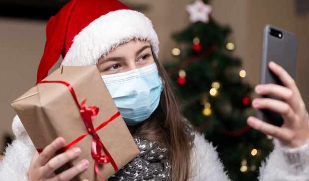 Auguri di Natale in linea. Una giovane ragazza con un cappello di Babbo Natale e una maschera medica parla utilizzando lo smartphone per videochiamare amici e genitori. Natale durante il coronavirus