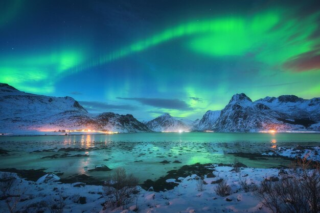 Aurora boreale sulla costa del mare montagne innevate e luci della città di notte Aurora boreale nelle isole Lofoten Norvegia Cielo stellato con luci polari Paesaggio invernale con aurora riflessa nell'acqua