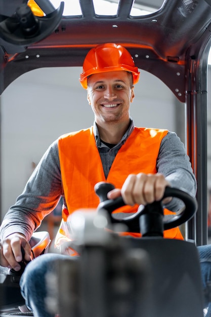 Autista di fabbrica sorridente che indossa casco e abbigliamento da lavoro che guida un carrello elevatore che lavora in magazzino