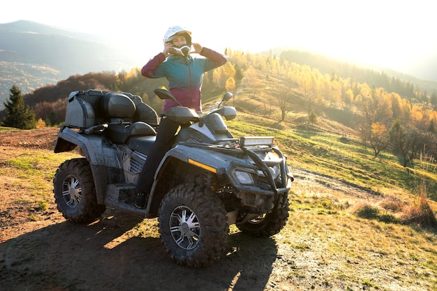 Autista donna felice in casco protettivo godendo di guida estrema su moto quad ATV in montagne estive al tramonto.