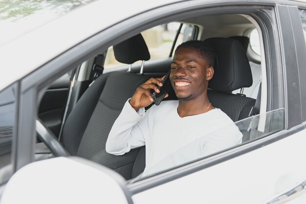 Autista maschio afroamericano sorridente felice seduto dietro il volante a guida autonoma di un'auto elettrica autonoma moderna. Il ragazzo felice tiene il telefono e sorride alla macchina fotografica in un'auto elettrica moderna