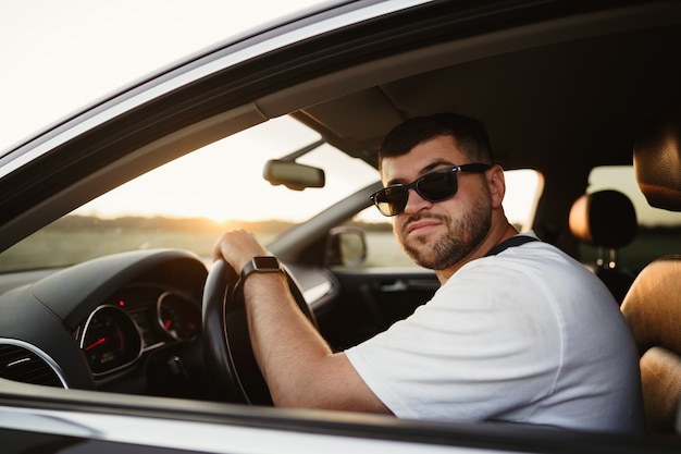 Autista uomo barbuto in occhiali da sole seduto nella sua auto e guardando la telecamera