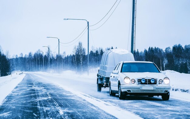 Auto con rimorchio in neve invernale su strada in Finlandia. Veicolo automatico con trasportatore trasportatore Carrier su strada privata. Logistica dei trasporti europea al trasporto di lavoro di trasporto. Traino con conducente in autostrada.