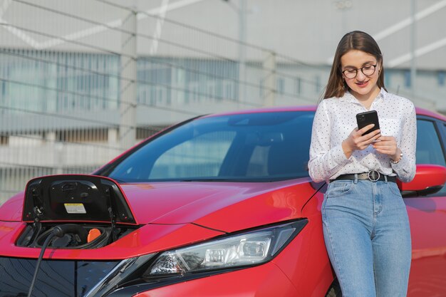 Auto ecologica collegata e ricarica batterie. La ragazza che per mezzo dello smartphone e l'attesa dell'alimentazione elettrica si collegano ai veicoli elettrici per caricare la batteria in automobile