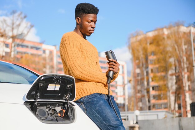 Auto elettriche, concept EV, carburante ecologico. Ritratto di giovane uomo di colore sorridente, che ricarica la sua moderna auto elettrica di lusso