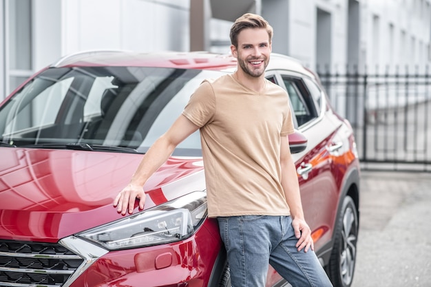 Auto rossa. Uomo sorridente in maglietta e jeans appoggiato su una nuova auto rossa scintillante all'aperto in città durante il giorno