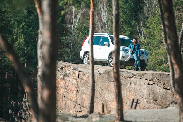 Automobile bianca del suv alla scogliera rocciosa giovane donna vicino ad essa vista attraverso gli alberi