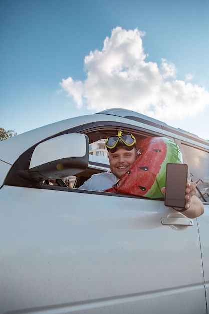 Automobile felice dell'uomo del conducente piena di roba di vacanza che tiene il telefono con lo schermo bianco