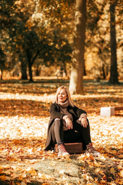 Autunno dorato. Elegante bionda in cappotto nero e maglione di lana con ampio colletto posa seduto su una vecchia valigia nella foresta autunnale su foglie cadute. Divertimento durante l'escursione nella stagione autunnale.